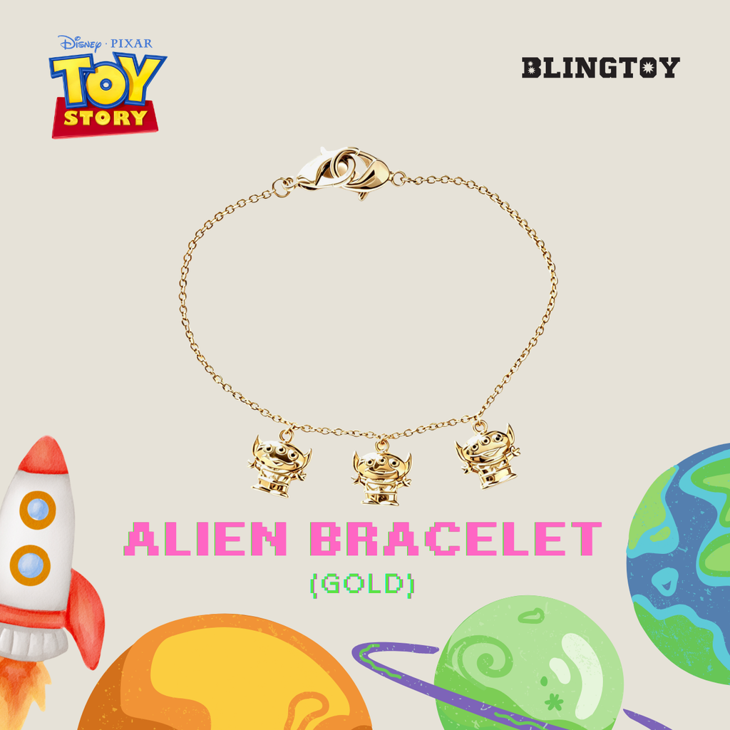 A gold-tone Alien bracelet featuring three dangling Alien figures on a delicate chain, presented on a beige background with Toy Story branding, planets, and a rocket illustration.